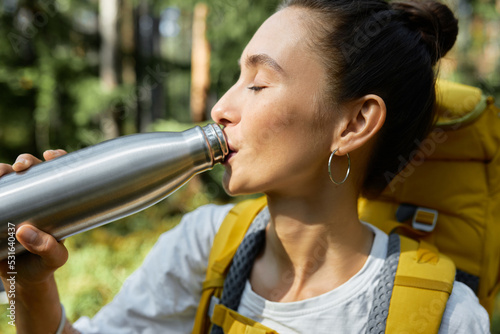 Close up of thirsty woman backpacker drinking fresh water or tea during stop in trekking in wild nature, enjoying drink with closed eyes, carrying rucksack on her shoulders with forest on background photo