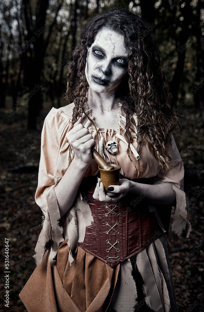 Halloween (Samhain) theme: scary gloomy voodoo witch with mortar and pestle. Portrait of evil sorceress in dark grove. Zombie woman (undead)