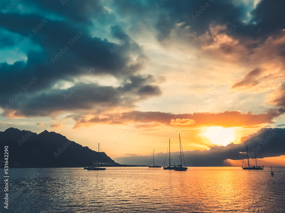 Majestic yacht tour travel landscape. Dramatic nature sunset in tropical island lagoon. Yachts silhouette against bright orange sun and blue clouds sky. Wonderful picturesque scene. French Polynesia