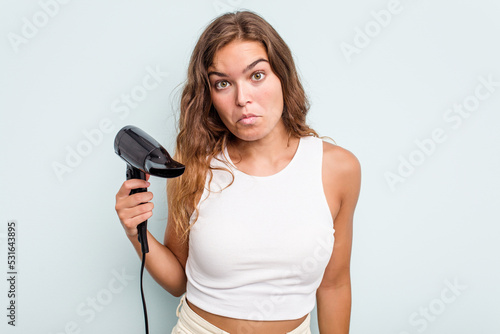 Young caucasian woman holding a hairdryer isolated on blue background shrugs shoulders and open eyes confused.