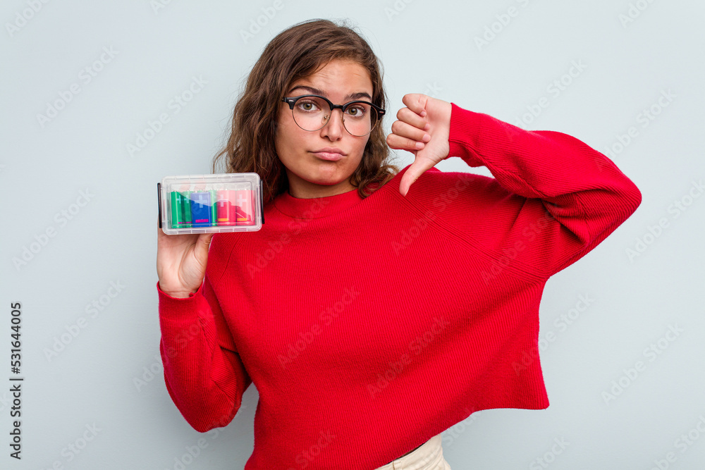 Naklejka premium Young caucasian woman holding battery box isolated on blue background showing a dislike gesture, thumbs down. Disagreement concept.