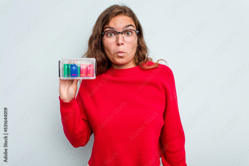 Young caucasian woman holding battery box isolated on blue background shrugs shoulders and open eyes confused. Young caucasian woman holding battery box isolated on blue background shrugs shoulders and open eyes confused.