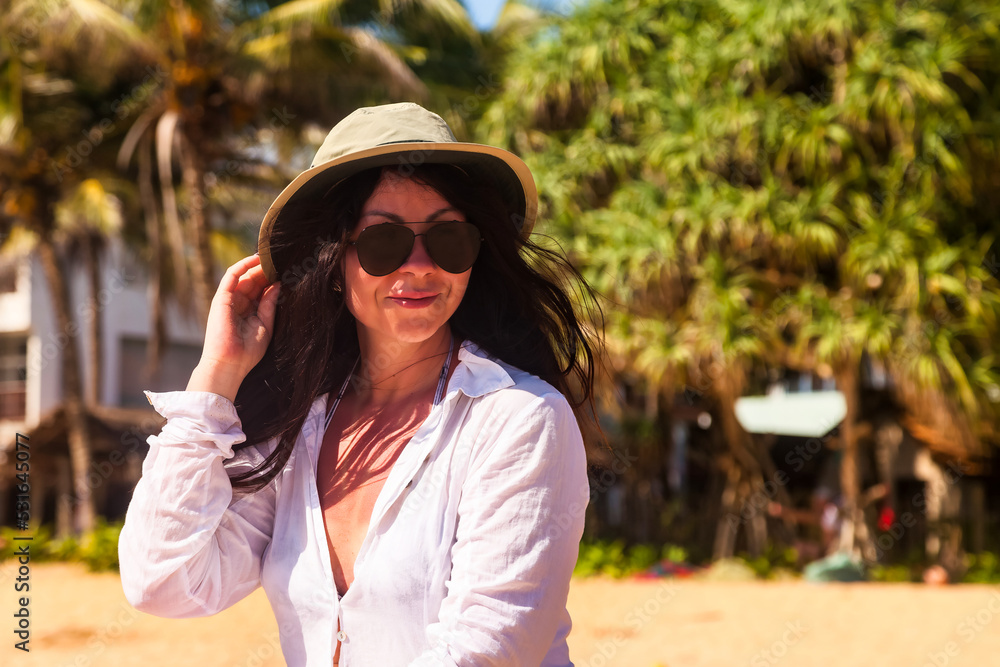 Portrait adult woman in white beachwear, hat and sunglasses smiling, looking away, sitting on tropical sandy beach at palm trees background. Travel tourism vacation concept. Copy text space