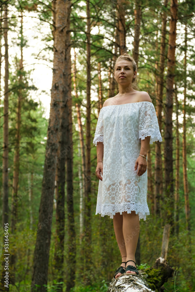 A young woman in a forest in a white dress stands on a tree trunk and looks into the distance. Vertical photo.