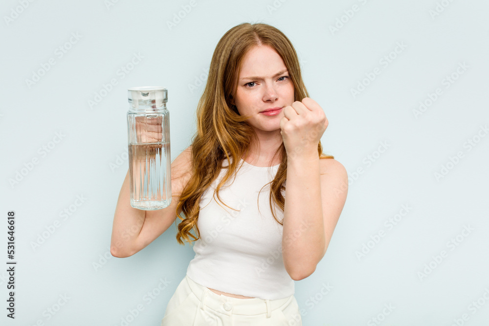 Young caucasian woman holding jar of water isolated on blue background showing fist to camera, aggressive facial expression. Young caucasian woman holding jar of water isolated on blue background showing fist to camera, aggressive facial expression.