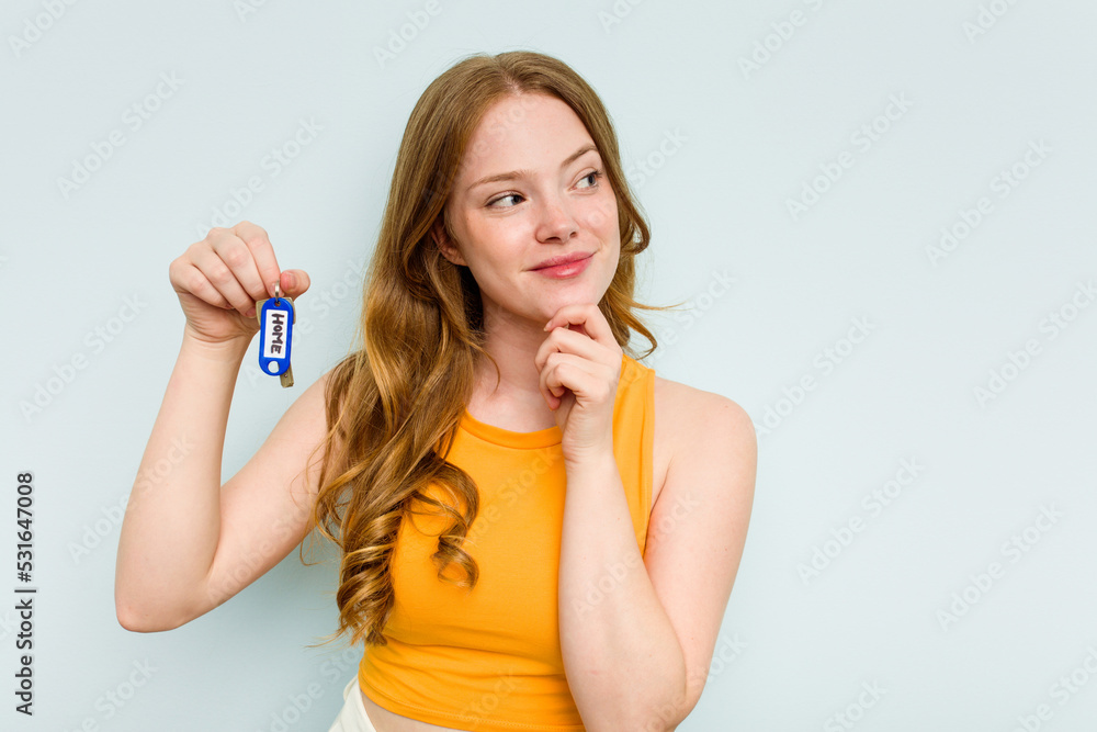 Fototapeta premium Young caucasian woman holding home keys isolated on blue background looking sideways with doubtful and skeptical expression.