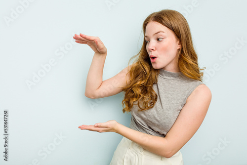 Young caucasian woman isolated on blue background shocked and amazed holding a copy space between hands.