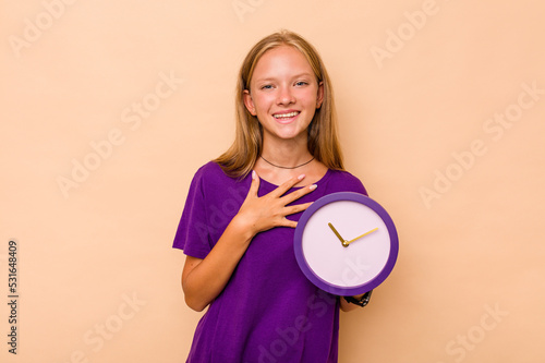 Little caucasian girl holding a clock isolated on beige background laughs out loudly keeping hand on chest.