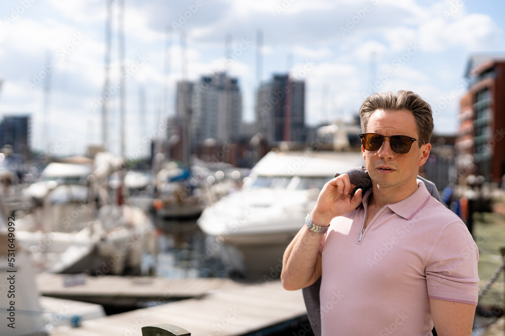 A handsome businessman in a casual grey suit in front of a busy marina with luxury yachts moored up A handsome businessman in a casual grey suit in front of a busy marina with luxury yachts moored up