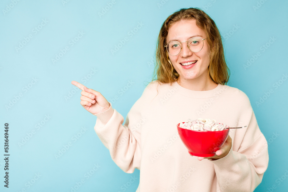 Young caucasian woman holding a bowl of cereals isolated on blue background smiling and pointing aside, showing something at blank space. Young caucasian woman holding a bowl of cereals isolated on blue background smiling and pointing aside, showing something at blank space.