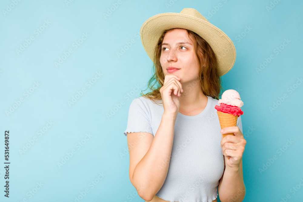 Young caucasian woman holding an ice cream isolated a blue background looking sideways with doubtful and skeptical expression.