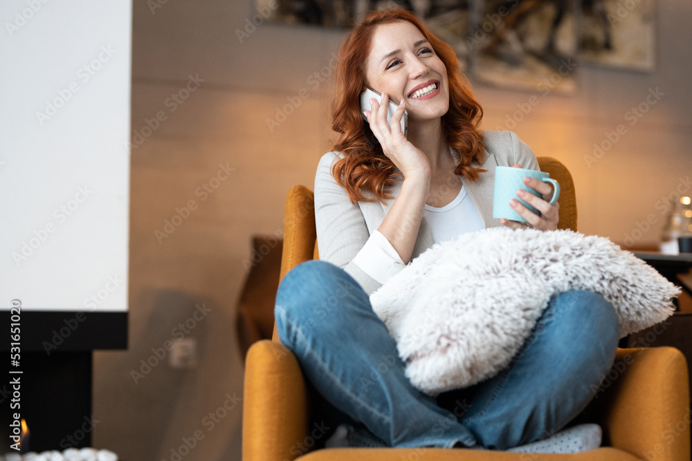 Beautiful, young, smiling woman sitting in living room, enjoying her coffee and talking on phone Beautiful, young, smiling woman sitting in living room, enjoying her coffee and talking on phone