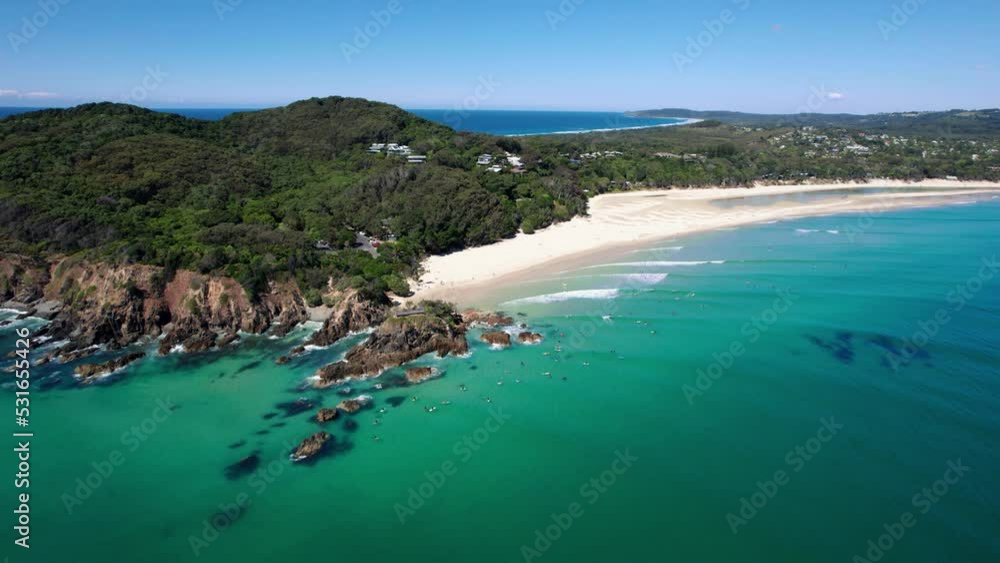 Drone orbits around The Pass in Byron Bay on a beautifully calm morning. Surfers surfing on small waves in crystal clear water. Beaches and green forest coastal backdrop. Aerial shot in 5.4K.