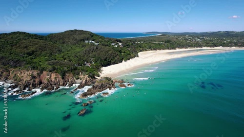 Drone orbits around The Pass in Byron Bay on a beautifully calm morning. Surfers surfing on small waves in crystal clear water. Green forest backdrop. Aerial shot in 5.4K. photo