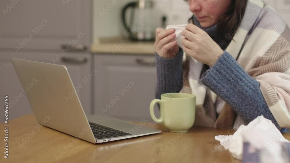 sick woman wrapped warm blanket sit kitchen table take paper napkin blow running nose working laptop looking computer screen. cold and flu illness season. heating shutdown high prices utilities sick woman wrapped warm blanket sit kitchen table take paper napkin blow running nose working laptop looking computer screen. cold and flu illness season. heating shutdown high prices utilities