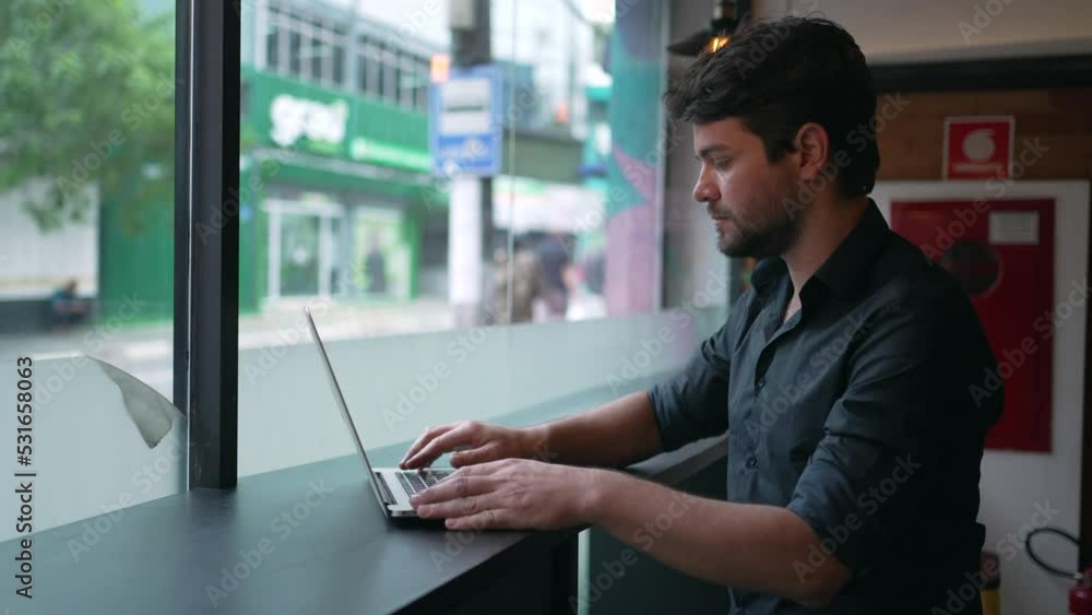 Man opening laptop seated at co working office by window overlooking city street sidewalk. Person works remotely Man opening laptop seated at co working office by window overlooking city street sidewalk. Person works remotely