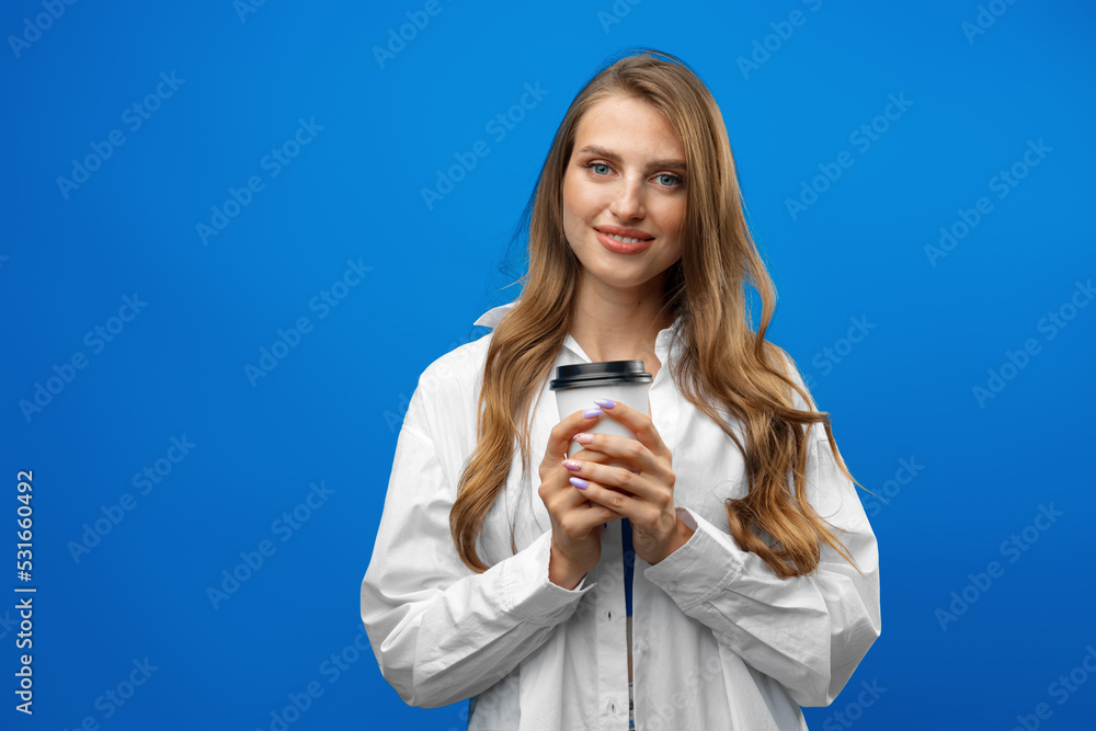 Beautiful attractive young lady holding takeaway paper coffee cup against blue background in studio