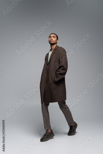 Foto full length of young african american man in autumnal coat looking at camera and posing with hand in pocket on grey