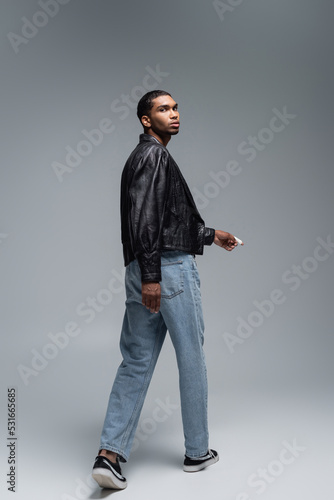 full length of young african american man in leather jacket walking on grey.