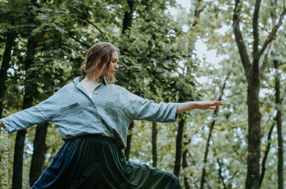 Woman doing yoga warrior II pose in oversized linen shirt with forest background Woman doing yoga warrior II pose in oversized linen shirt with forest background