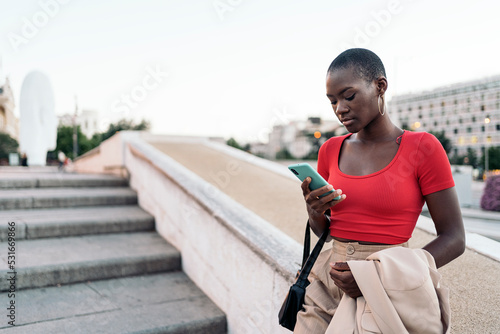 Young adult woman using her phone while holding her jacket and bag on outdoor stairs in the city
