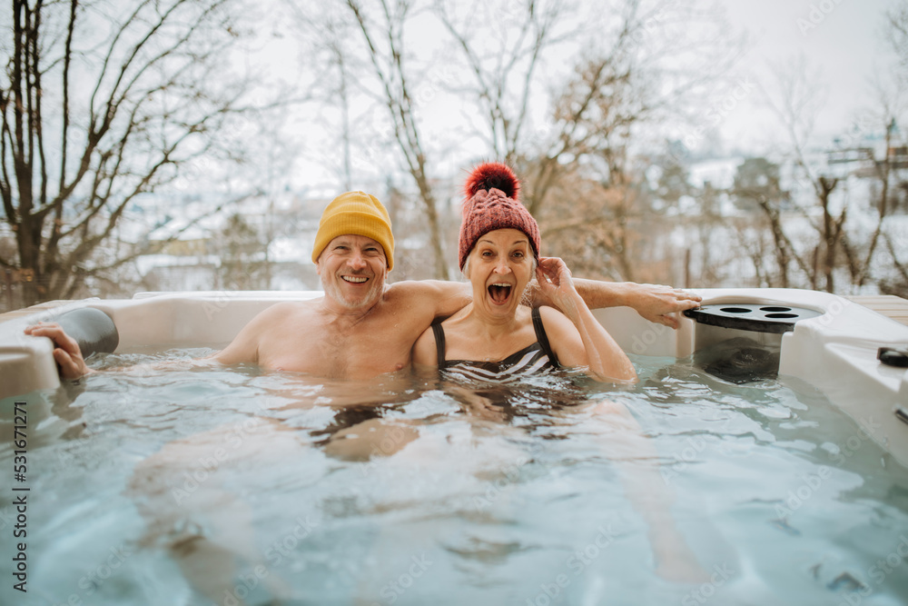 Senior couple in kintted cap enjoying together outdoor bathtub at their terrace during cold winter day.