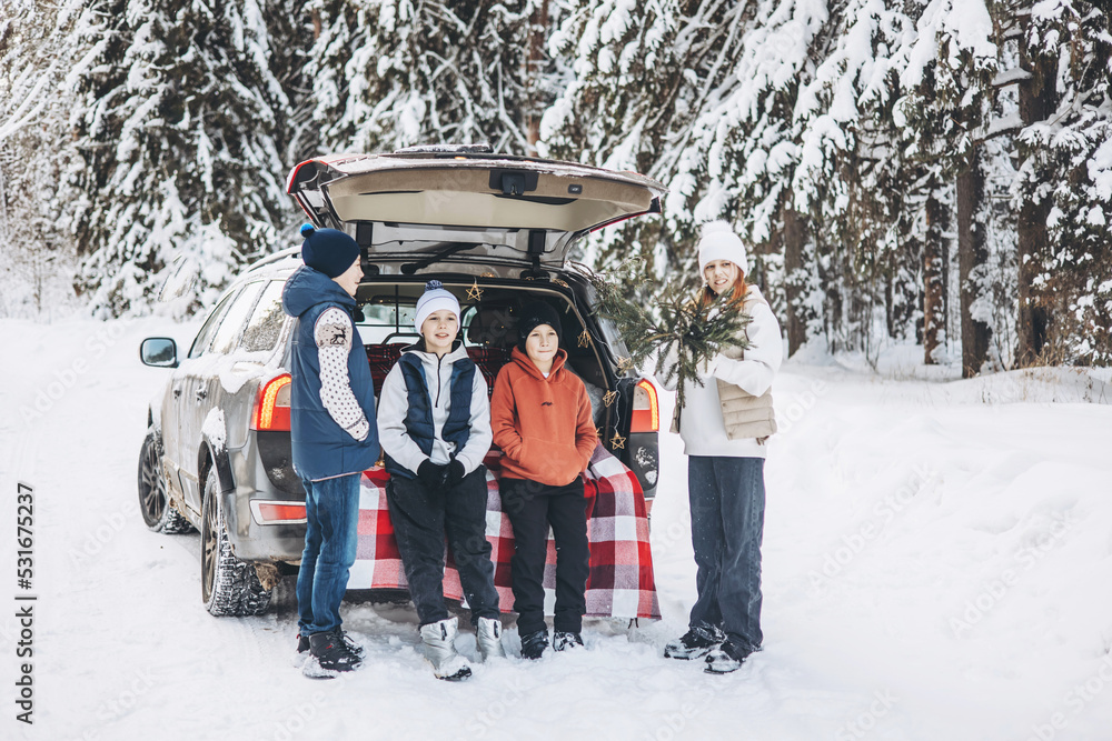 Four friends teenagers boys and girl with thermos sitting in trunk of car decorated for Christmas and New year. Winter picnic in snowy forest. Road trip and local travel. Friends hanging out together