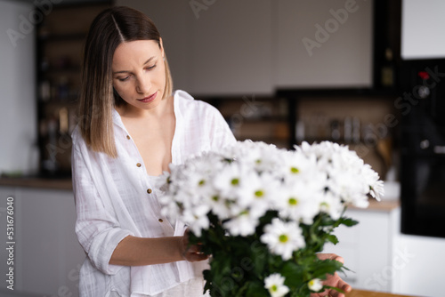 Happy and joyful young woman in white arranging white flowers at home in the kitchen