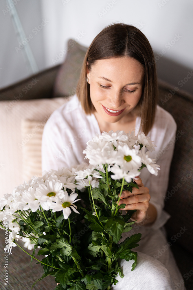 Good looking Caucasian woman enjoy flowers, she is happy to get a fresh bouquet of white chrysanthemum while relaxing on the sofa Good looking Caucasian woman enjoy flowers, she is happy to get a fresh bouquet of white chrysanthemum while relaxing on the sofa