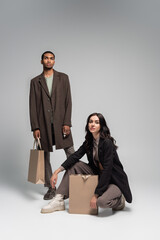 full length of stylish interracial couple in autumnal outfits posing near paper bags on grey.