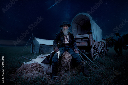 Fotografia Oldest smart cowboy man wearing western style suite with cowboy hat holding gun on hand sit on haystack with horse carrier and tent is vintage 1800s life style concept