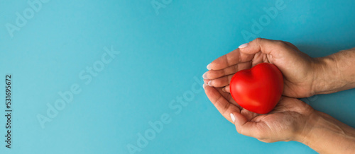 Fototapeta Naklejka Na Ścianę i Meble -  Hard hands hold a red heart on a blue background, banner, copyspace on the left. The concept of a woman's health, mom's love, mom's heart, loving parent giving care. Healthy heart