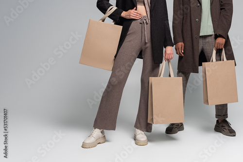 partial view of stylish interracial couple in autumnal outfits holding shopping bags on grey.