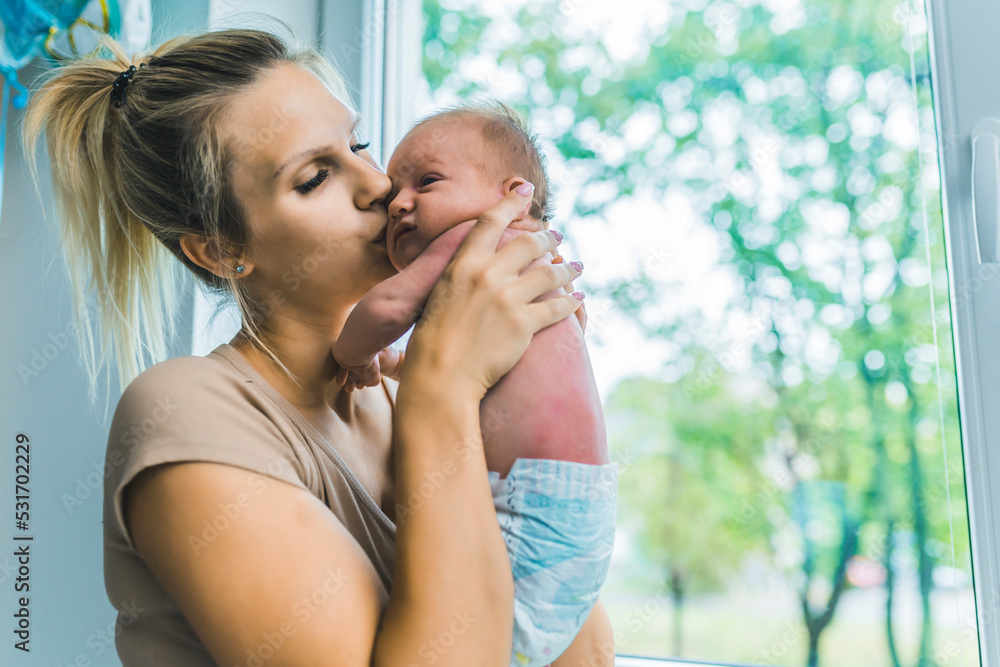 White blond woman standing in front of window holding up her newborn baby kissing it on the cheek. Happy mother. Horizontal indoor shot . High quality photo