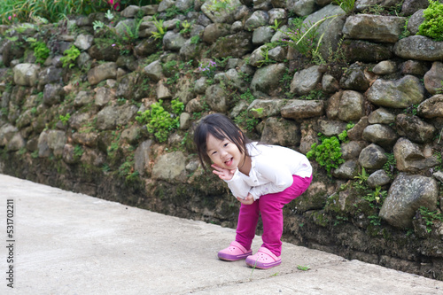 An Asian kid in a white shirt and purple pants in action with a smiley face in natural daylight. An Asian girl kid with rocks in the background.