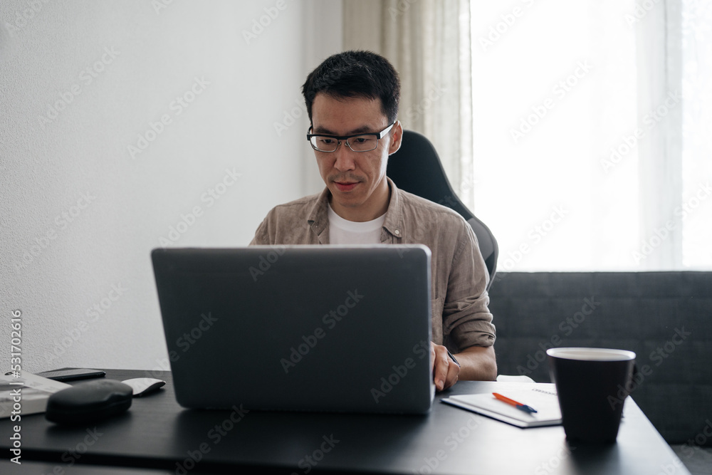 Adult asian man sitting at table using laptop at home. Remote job, freelance working concept. Adult asian man sitting at table using laptop at home. Remote job, freelance working concept.