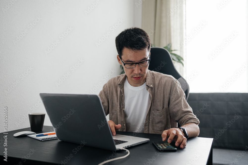 Happy adult asian man wearing glasses using laptop talking on smartphone at home. Remote job, freelance working concept. Happy adult asian man wearing glasses using laptop talking on smartphone at home. Remote job, freelance working concept.