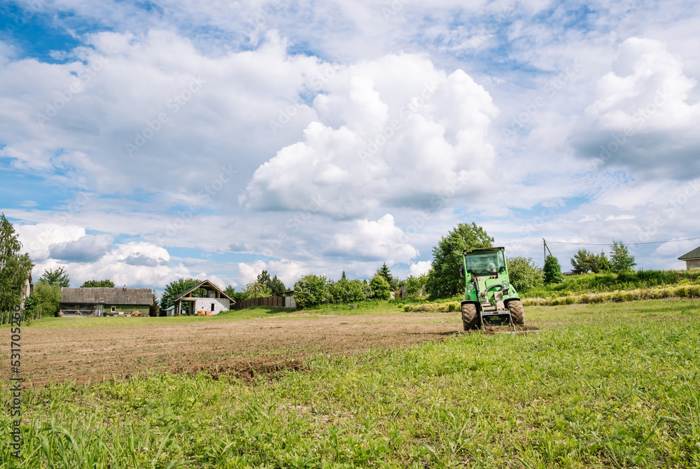 Fototapeta premium A green mini skid steer loader clear the construction site. Land work by the territory improvement. Small tractor with a ground leveler for moving soil, turf. Machine for agriculture work. Copy space