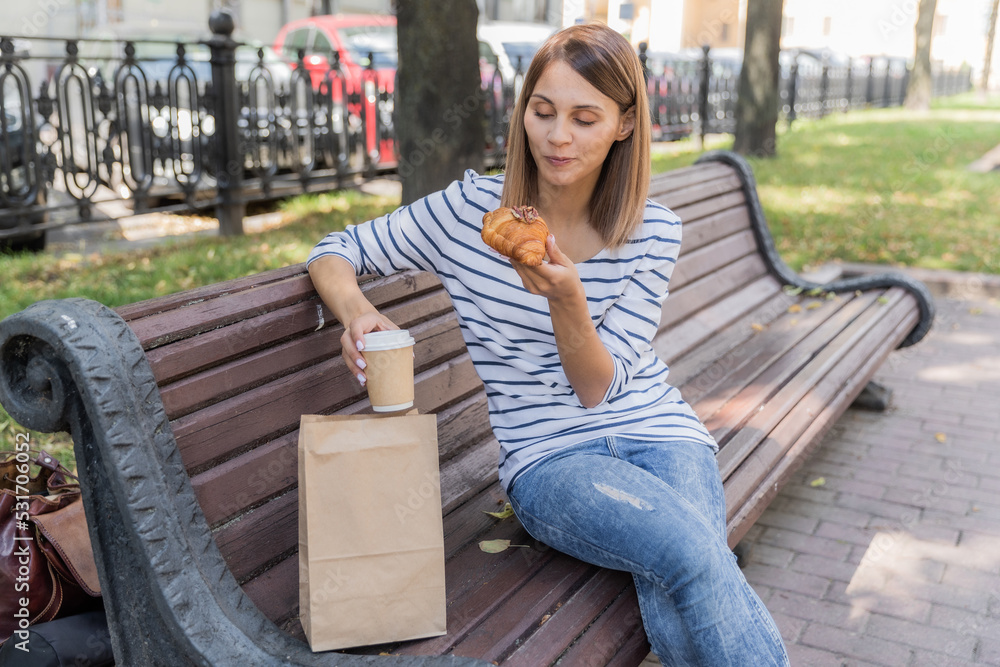 The girl is sitting on a bench in the park. Girl drinking coffee from a paper cup with plastic cap. Girl eating croissant. Sunny day in park. Vacation in park. Perspective in photo.