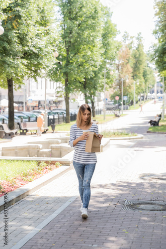 A girl in jeans and a vest walks around the square and drinks coffee with a croissant. Coffee in a paper cup and a croissant in a paper bag. Sunny day in the park with coffee and a croissant.
