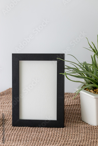 Blank vertical black frame beside a houseplant, painting or artwork display on brown knitted cloth
