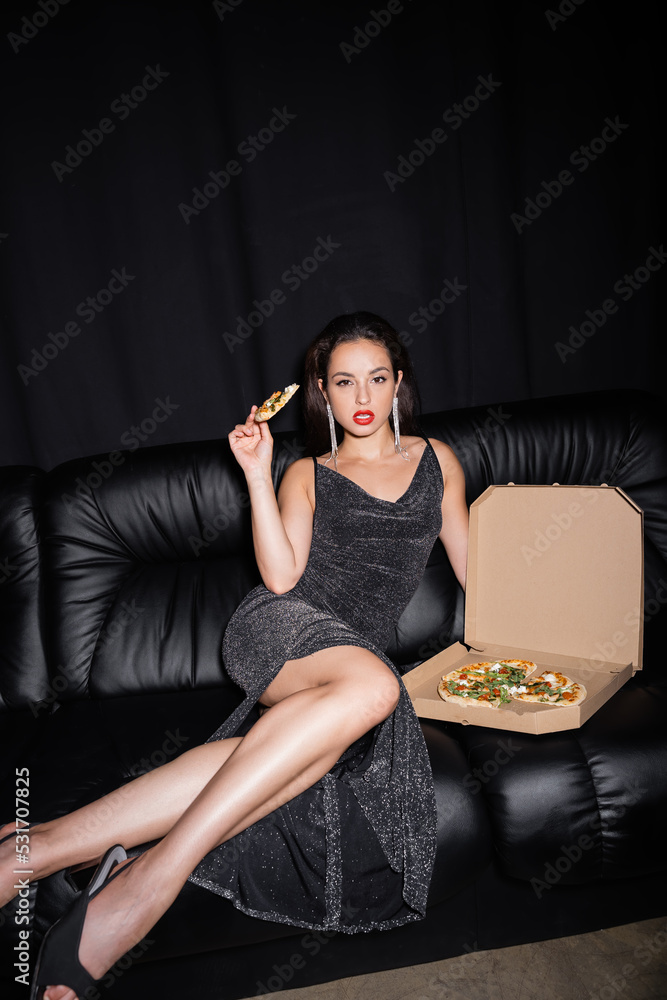 Naklejka premium elegant woman in lurex dress sitting on leather couch with pizza and looking at camera isolated on black.
