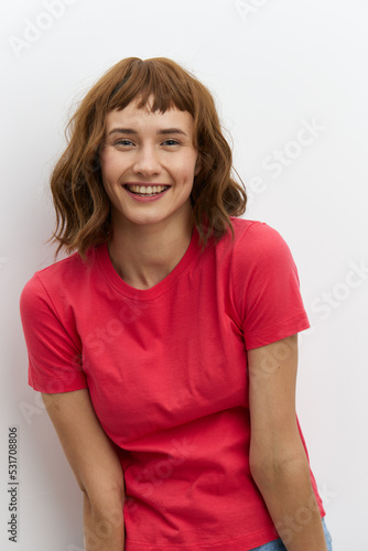 vertical photo of a beautiful, sweet woman standing on a light background in a red T-shirt and smiling pleasantly at the camera