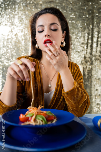 sexy woman in elegant dress touching lip near blurred plate with pasta on glitter background.