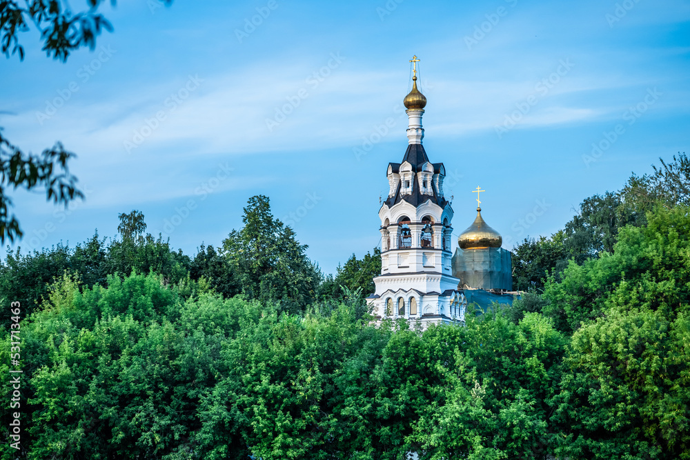 A beautiful church against the blue sky. The church is behind the trees. Colorful landscape. Ancient architecture. Green leaves. general plan