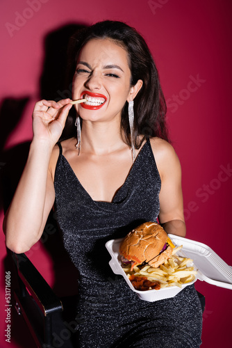 cheerful woman in black elegant dress eating french fries near burger in plastic container on red background.