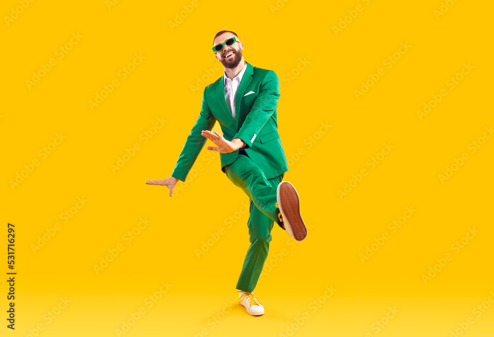 Obraz premium Funny young guy in a party suit dancing in a modern studio. Full body shot of a happy, cheerful, goofy man wearing a green suit and sunglasses dancing isolated on a bright yellow colour background