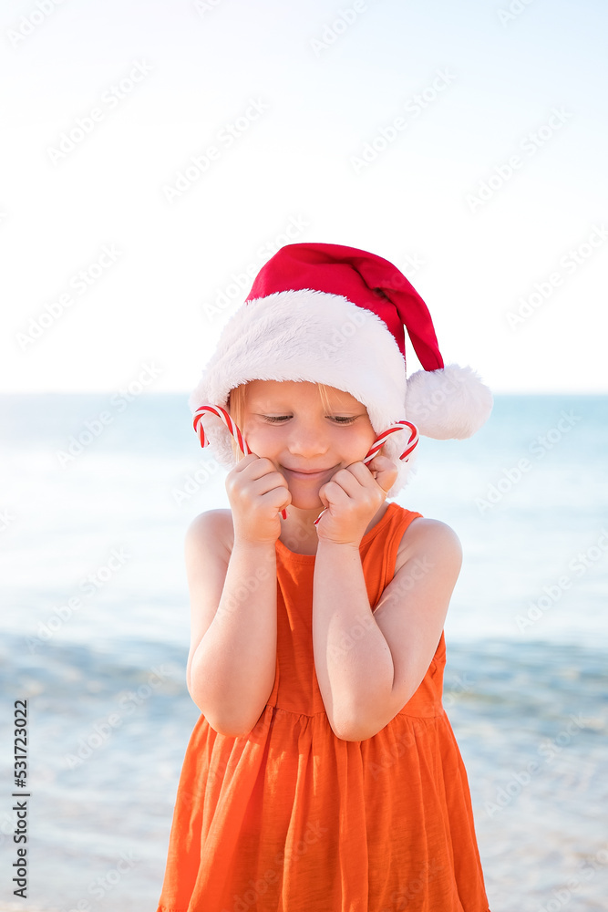 Obraz premium Girl in an orange dress, wearing a Santa Claus hat and holding Christmas lollipops on the beach is happy. Merry Christmas and Happy New Year.