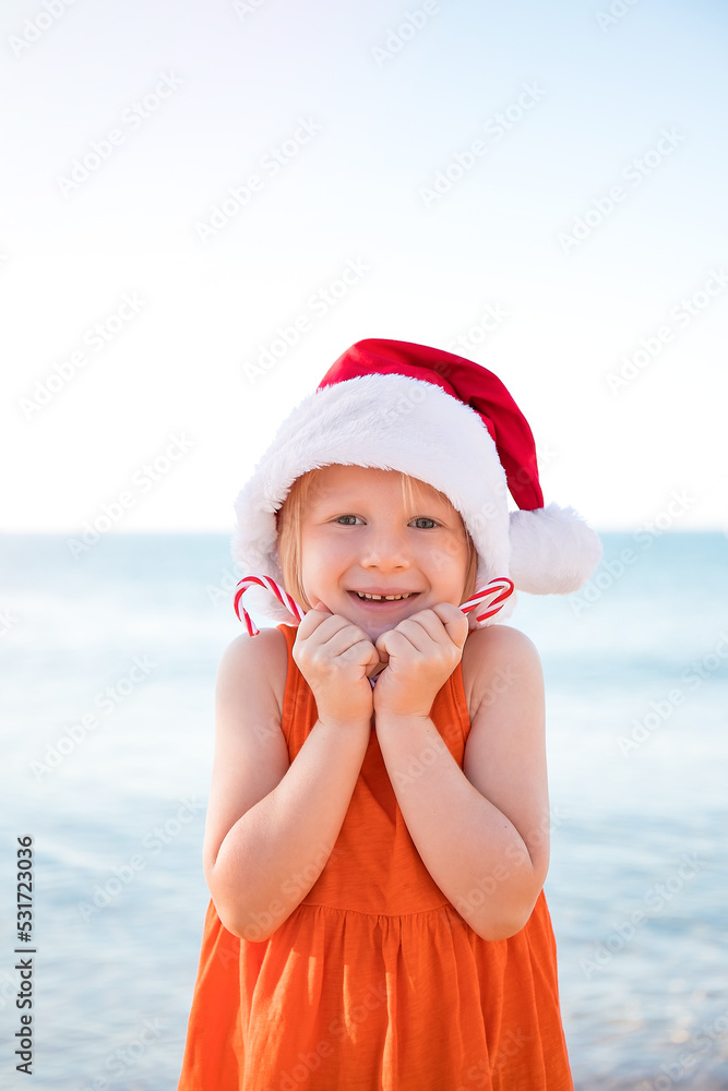Obraz premium Girl in an orange dress, wearing a Santa Claus hat and holding Christmas lollipops on the beach is happy. Merry Christmas and Happy New Year.