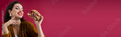 joyful and fashionable woman holding tasty burger isolated on red  banner.